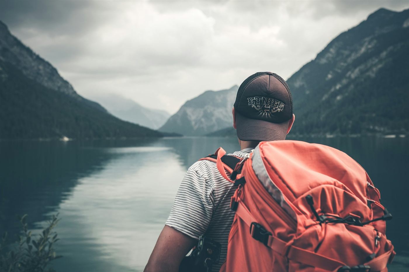 A traveler with a red backpack looking out over a calm mountain lake